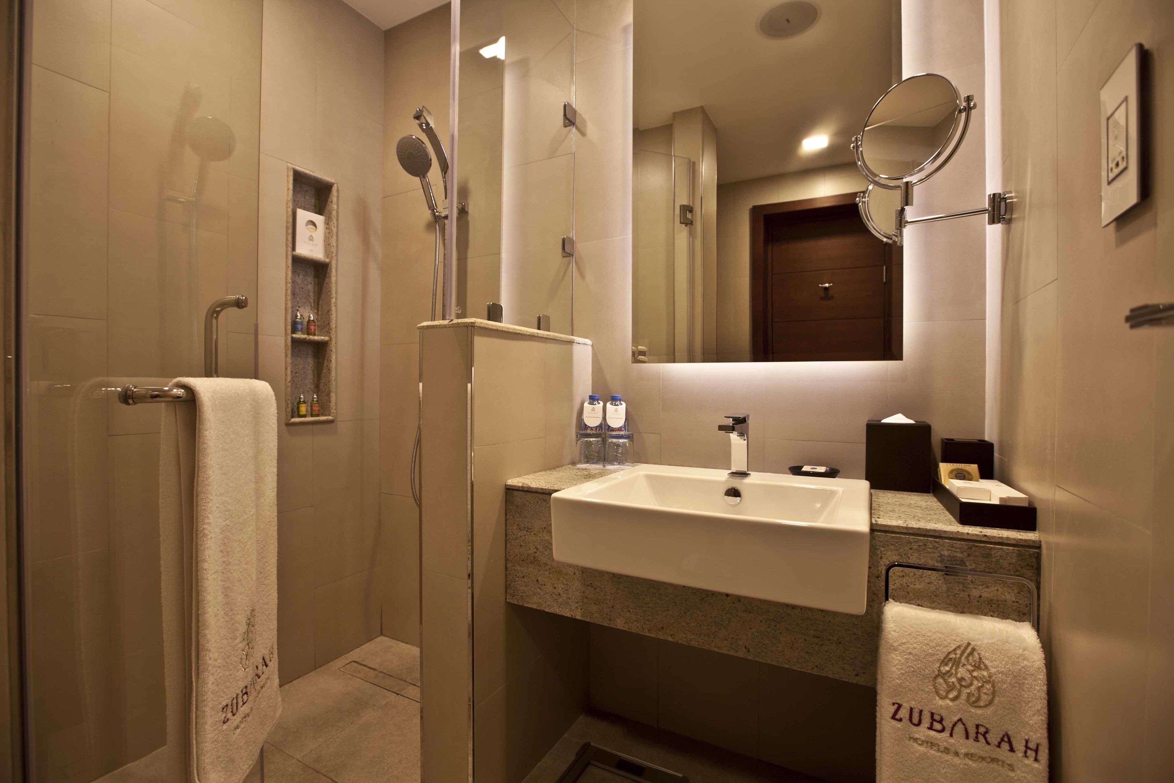 Une salle de bain moderne dotée d’une douche à parois de verre, d’un lavabo avec un miroir et de serviettes portant la marque « ZUBARAH ».