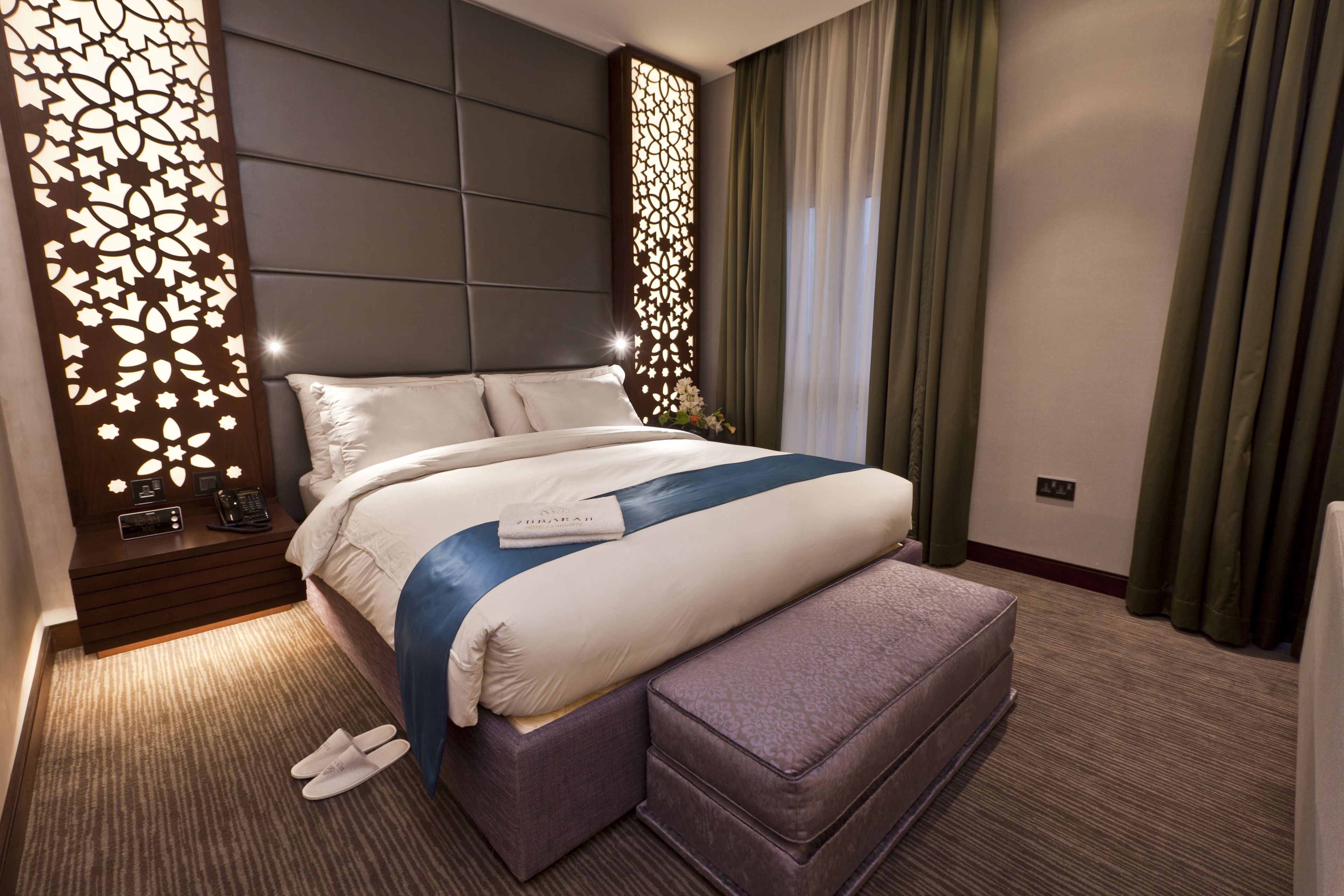 A modern hotel room with a large bed, a bedside table with a phone and a lamp, a decorative wall panel, and a view of the outside through a window with curtains.