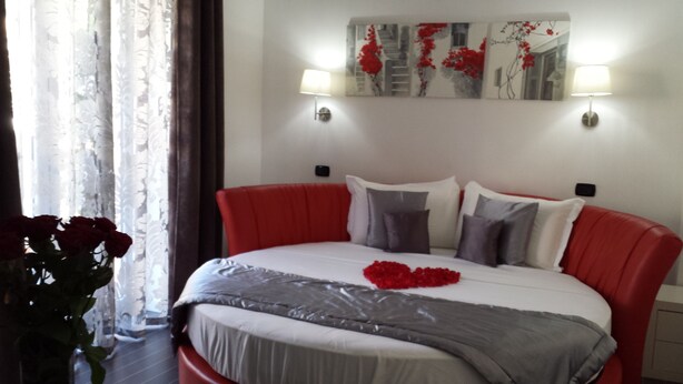 Una cama con cabecera roja, ropa de cama blanca y una manta gris. Hay dos lámparas de pared, un cuadro sobre la cama y un jarrón con flores rojas en el suelo.
