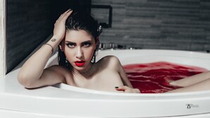Una persona en una bañera con agua roja, una pared oscura y una pulsera visible.