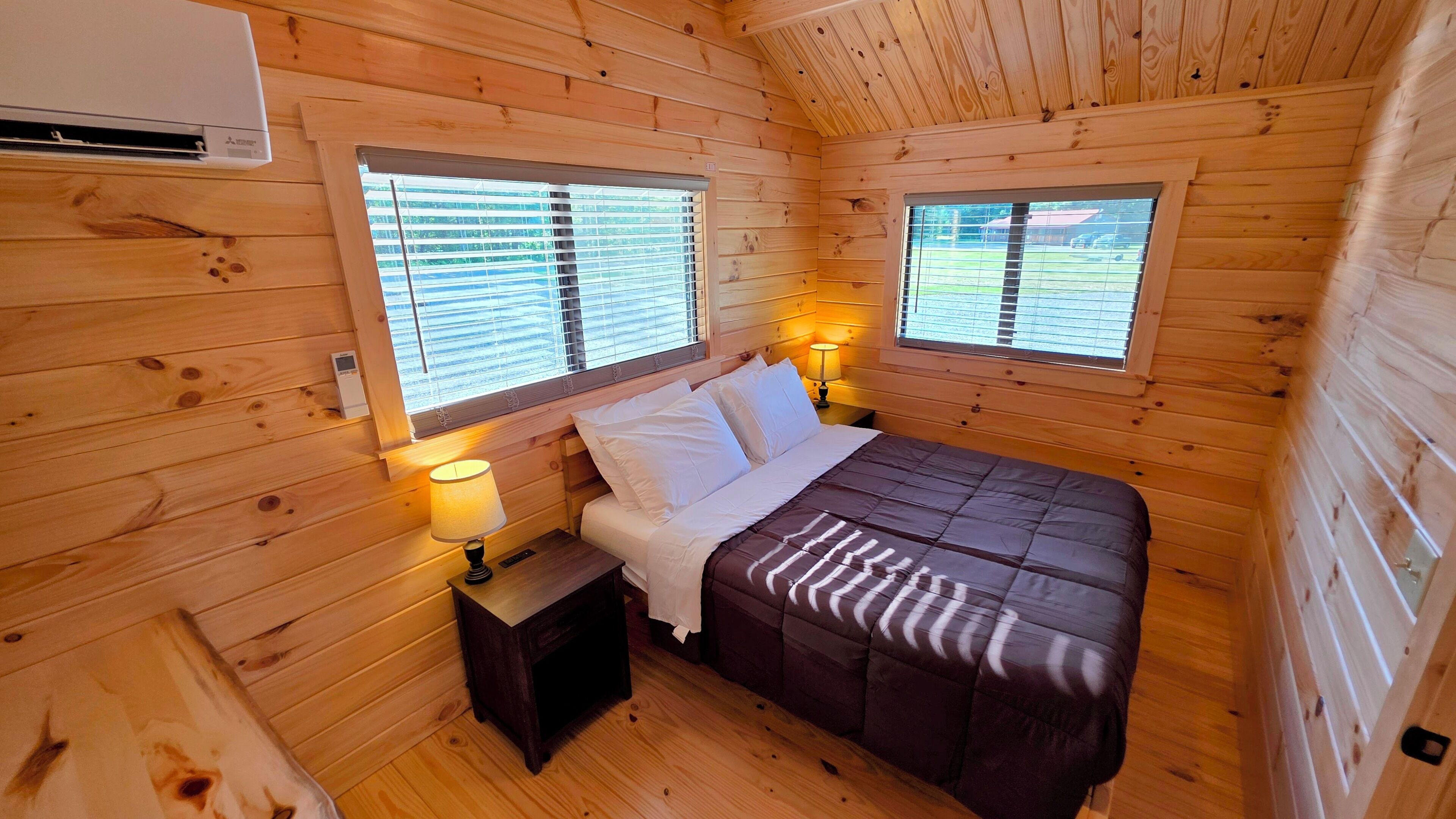 Quarto de cabana com uma cama, duas janelas, uma mesa de cabeceira e um abajur.