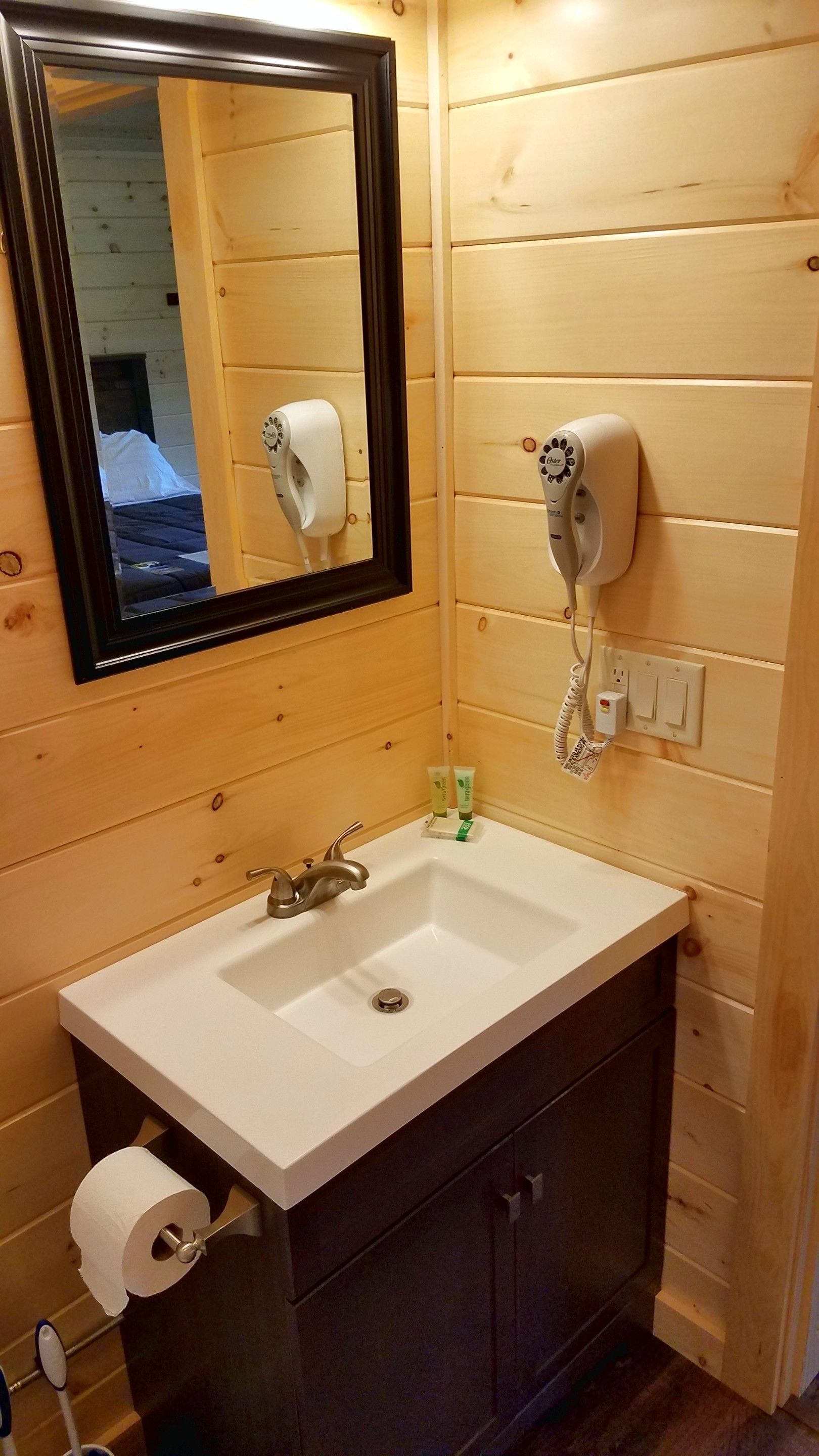 A wooden-paneled bathroom with a white sink, a mirror, a hairdryer, and a toilet paper holder.