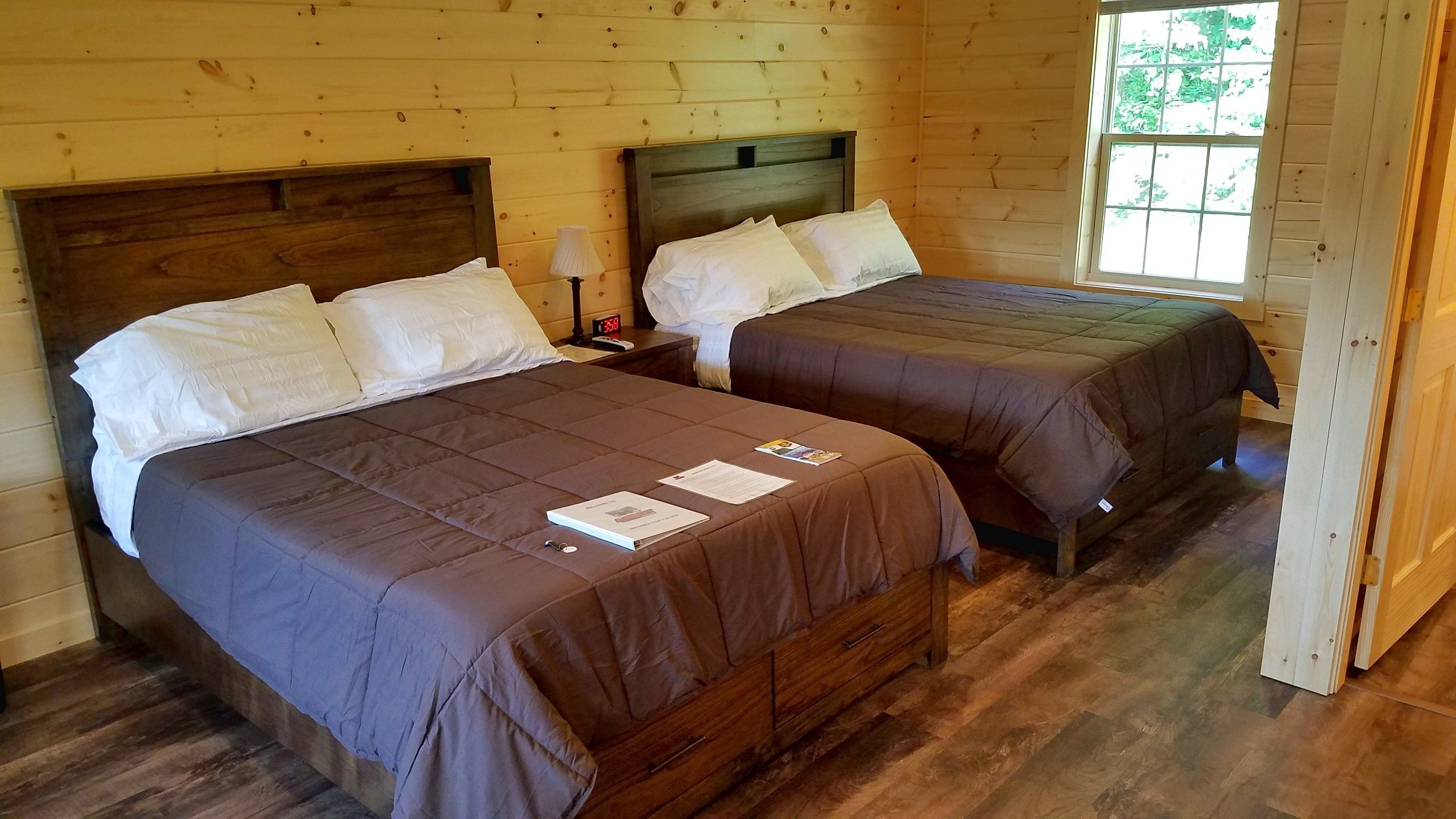 A wooden cabin-style room with two beds, each with a wooden headboard, a nightstand with a lamp, and a window with a view of greenery.