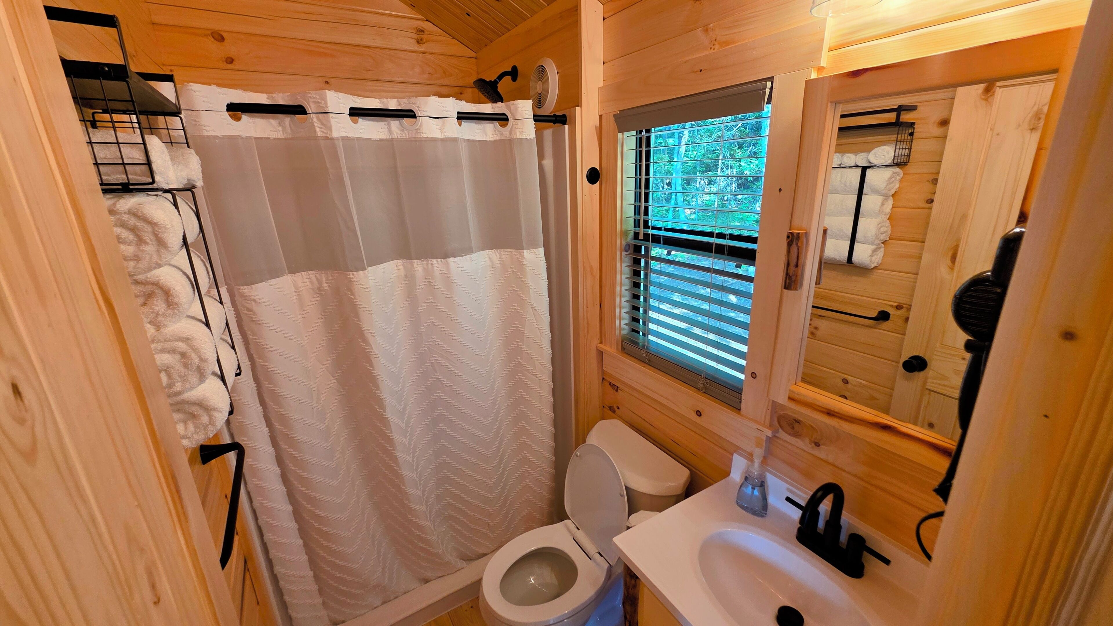 Een kleine houten badkamer met douchegordijn, toilet, wastafel en een raam met zonwering.