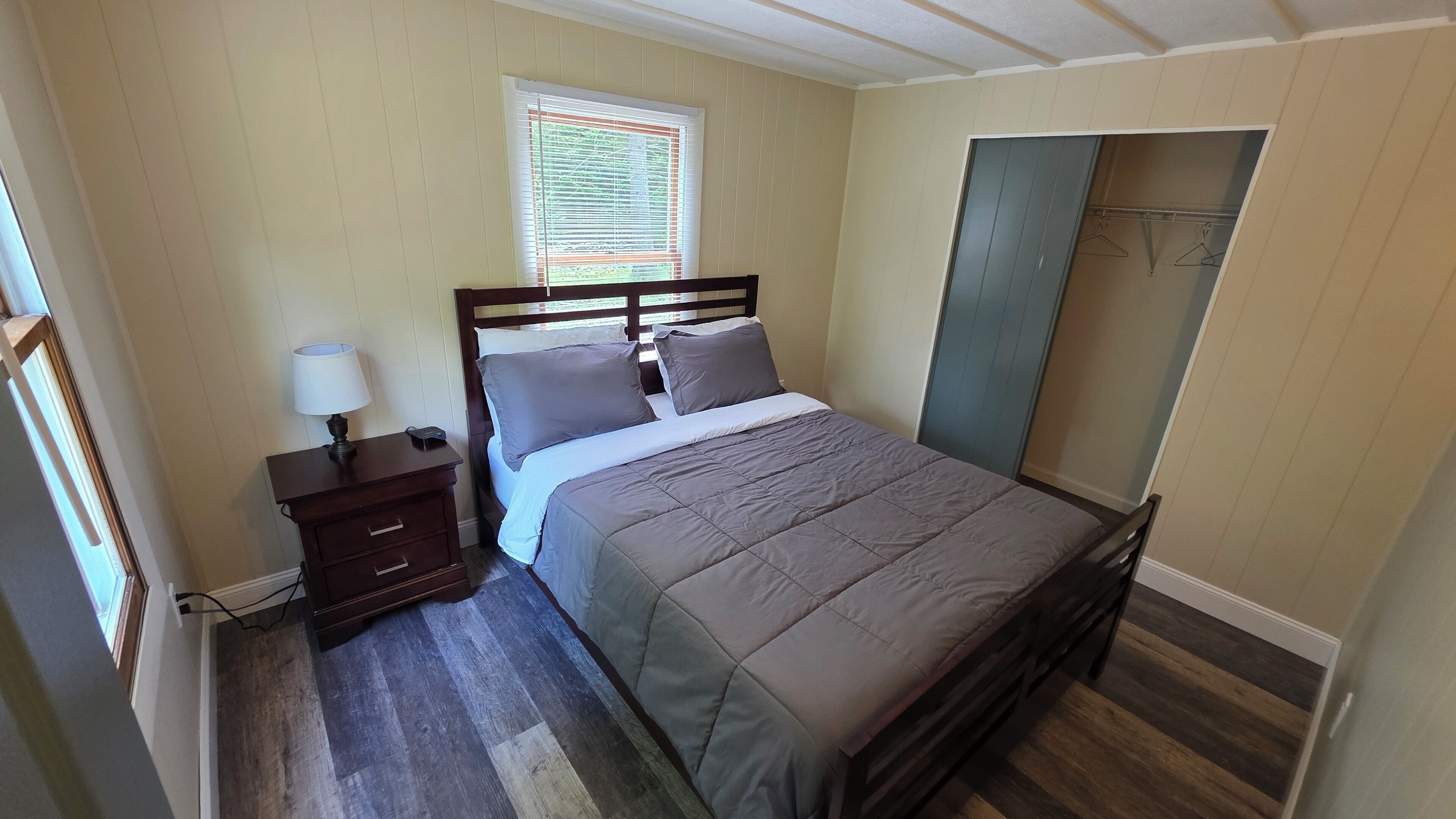 A bedroom with a bed, a nightstand, a lamp, a window with blinds, and a wardrobe.
