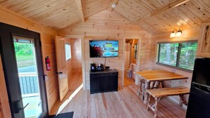 A cabin interior with a wooden ceiling, a television mounted on the wall, a small kitchen area with a table and bench, and a door leading outside. - Abbot Trailside Lodging (Abbot)