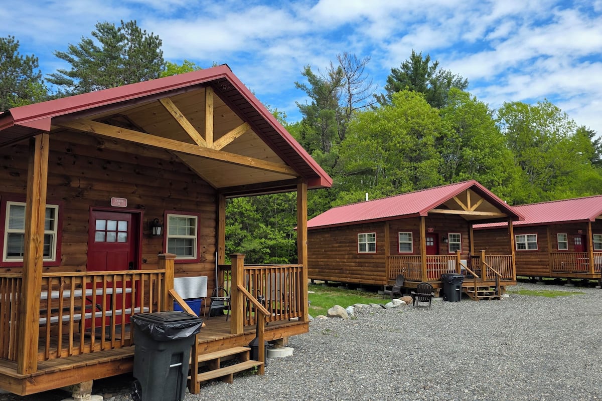 A row of log cabins with wooden decks and red roofs, surrounded by trees.