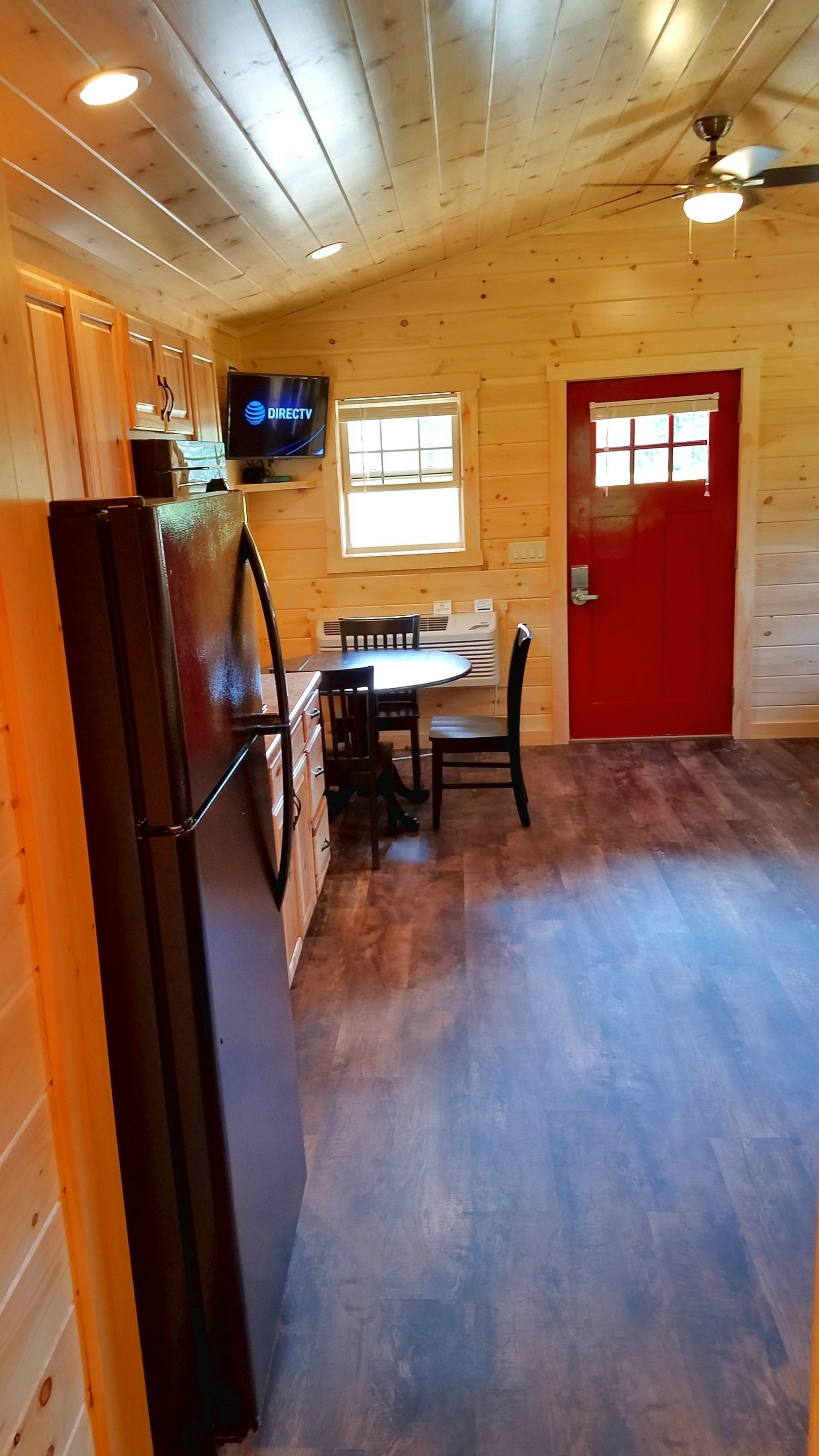 L’intérieur d’un chalet en bois comprend un coin cuisine équipé d’un réfrigérateur, une table à manger avec des chaises, un téléviseur fixé au mur et une porte rouge.