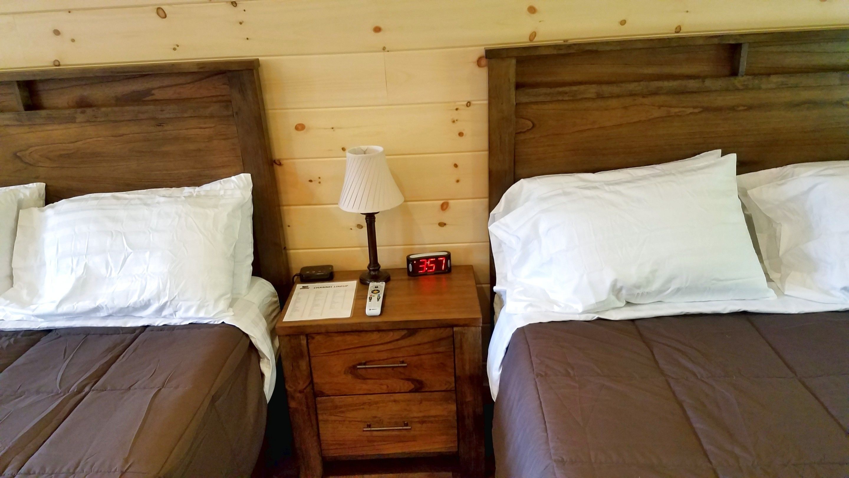 A double room with two wooden beds, white linens, a nightstand with a lamp, a digital clock, and a remote control.