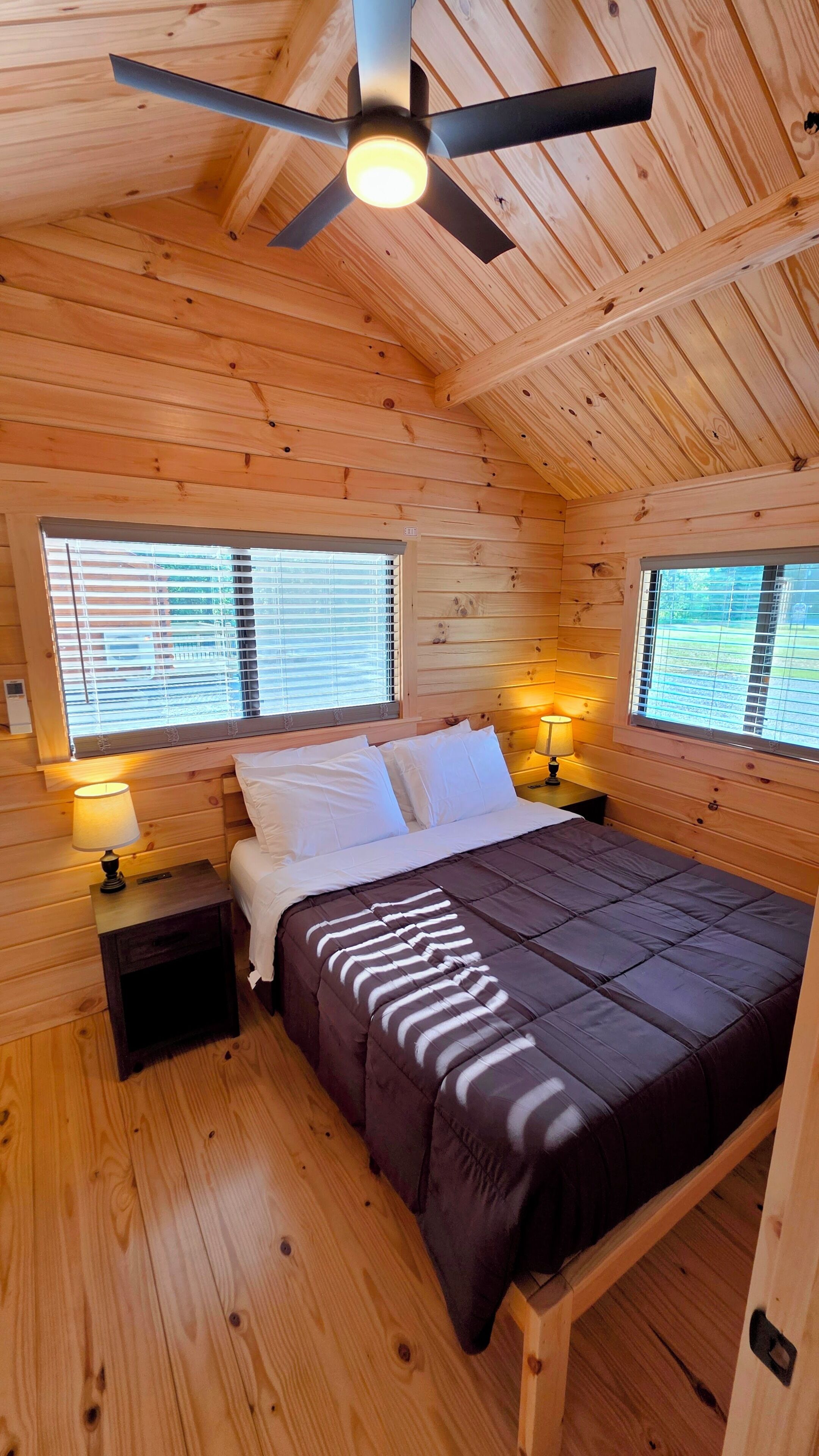 A wooden cabin bedroom with a bed, two bedside lamps, a ceiling fan, and windows with blinds.