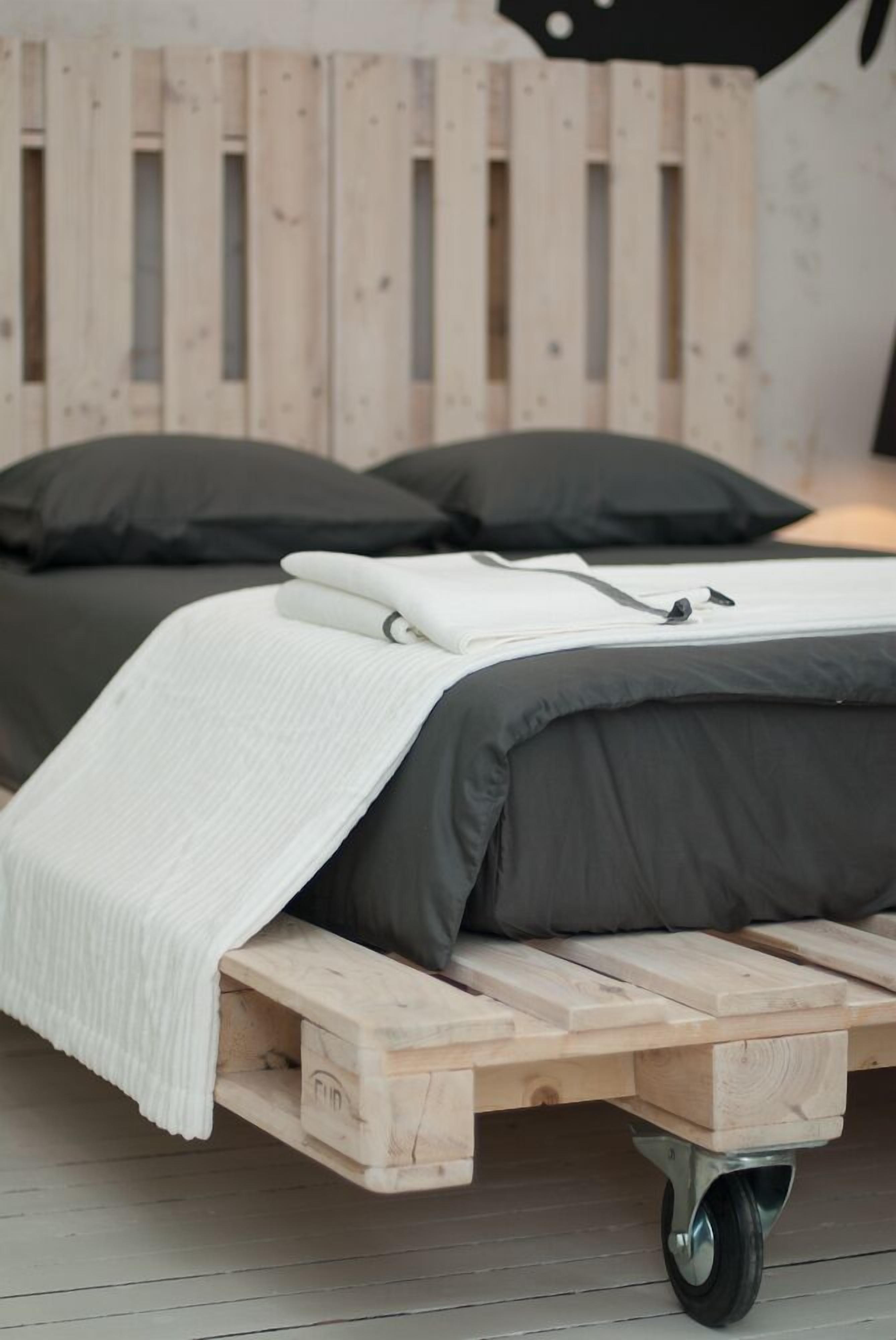 Ein Bett mit grau-weiß gestreifter Matratze, einer weißen Decke und zwei dunklen Kissen auf einem Holzboden mit Rollen.