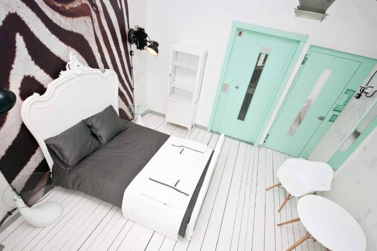 A bedroom with a zebra-patterned rug, a white bed with gray bedding, a white bedside table, a white lamp, a green door, and a white chair.