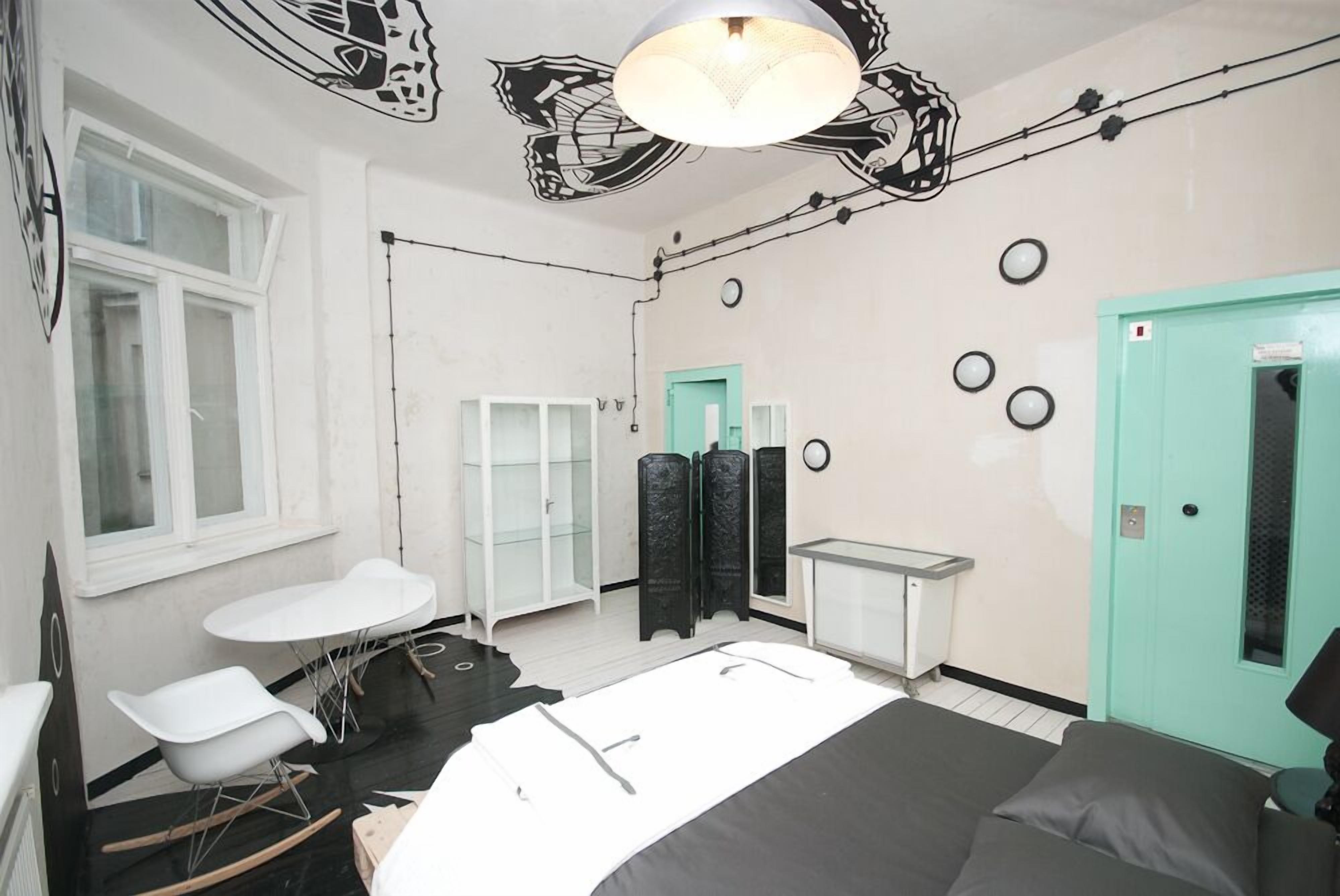 A room with a bed, a white table, a white chair, a teal door, and a black and white abstract wall design.