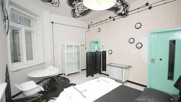 A room with a bed, a white table, a white chair, a teal door, and a black and white abstract wall design.