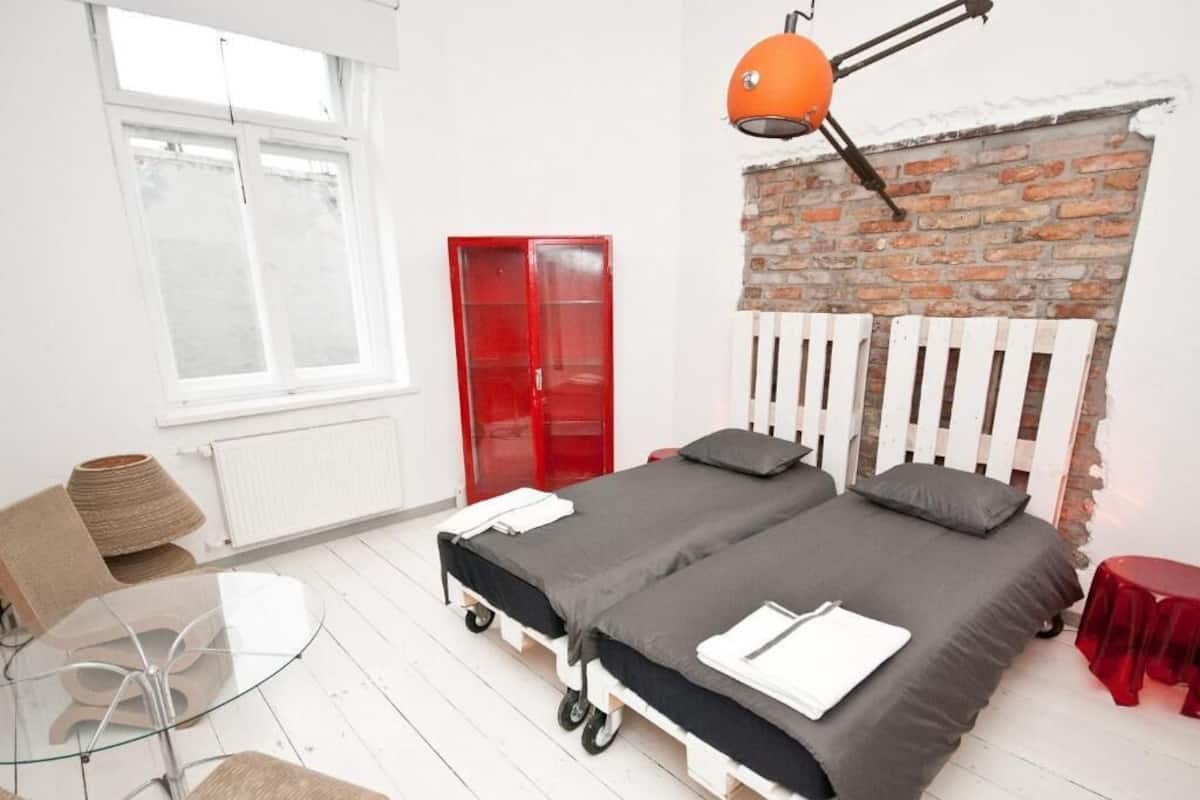 A bedroom with a double bed, a red wardrobe, a glass table, and a brick wall.
