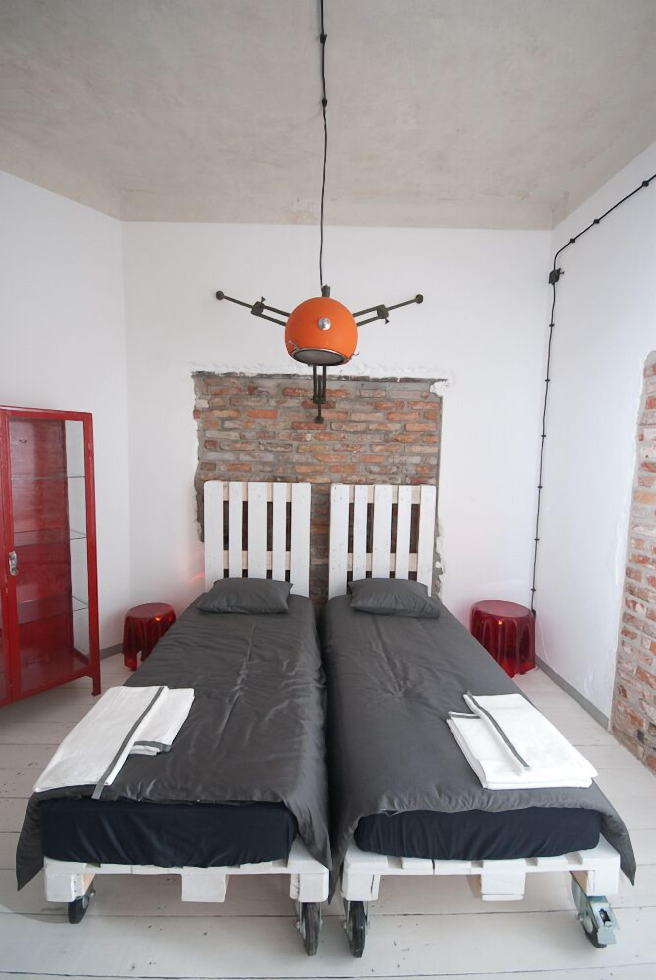 Ein Zimmer mit zwei Einzelbetten, einer Ziegelwand, einer roten Tür und einer hängenden, orangefarbenen Leuchte.