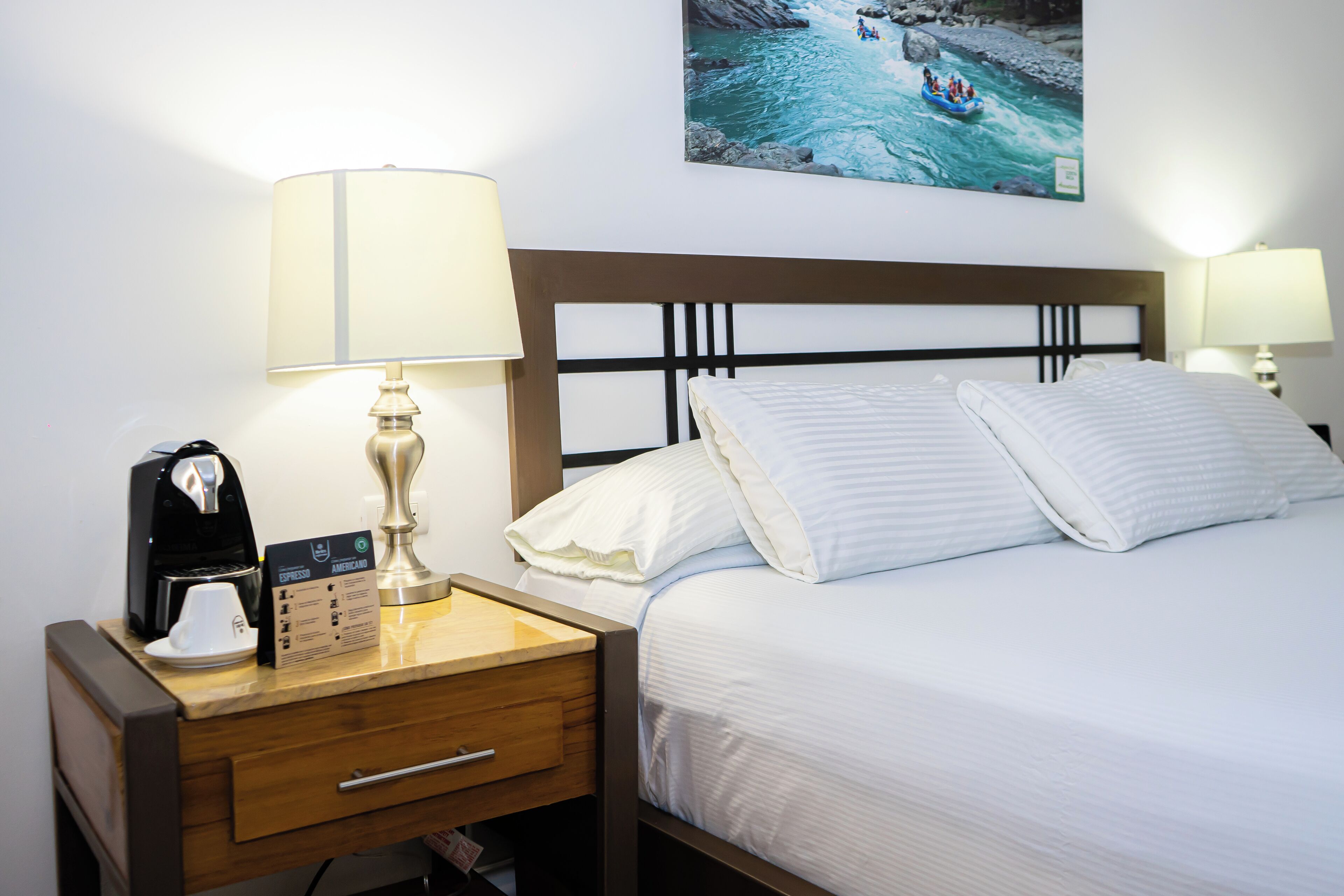 A neatly made bed with white linens, a wooden nightstand with a coffee maker, a lamp, and a framed picture of a river scene on the wall.