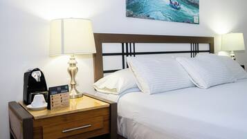 Un lit bien fait, avec du linge de lit blanc, une table de chevet en bois sur laquelle se trouvent une machine à café, une lampe et, au mur, un tableau représentant un paysage fluvial.