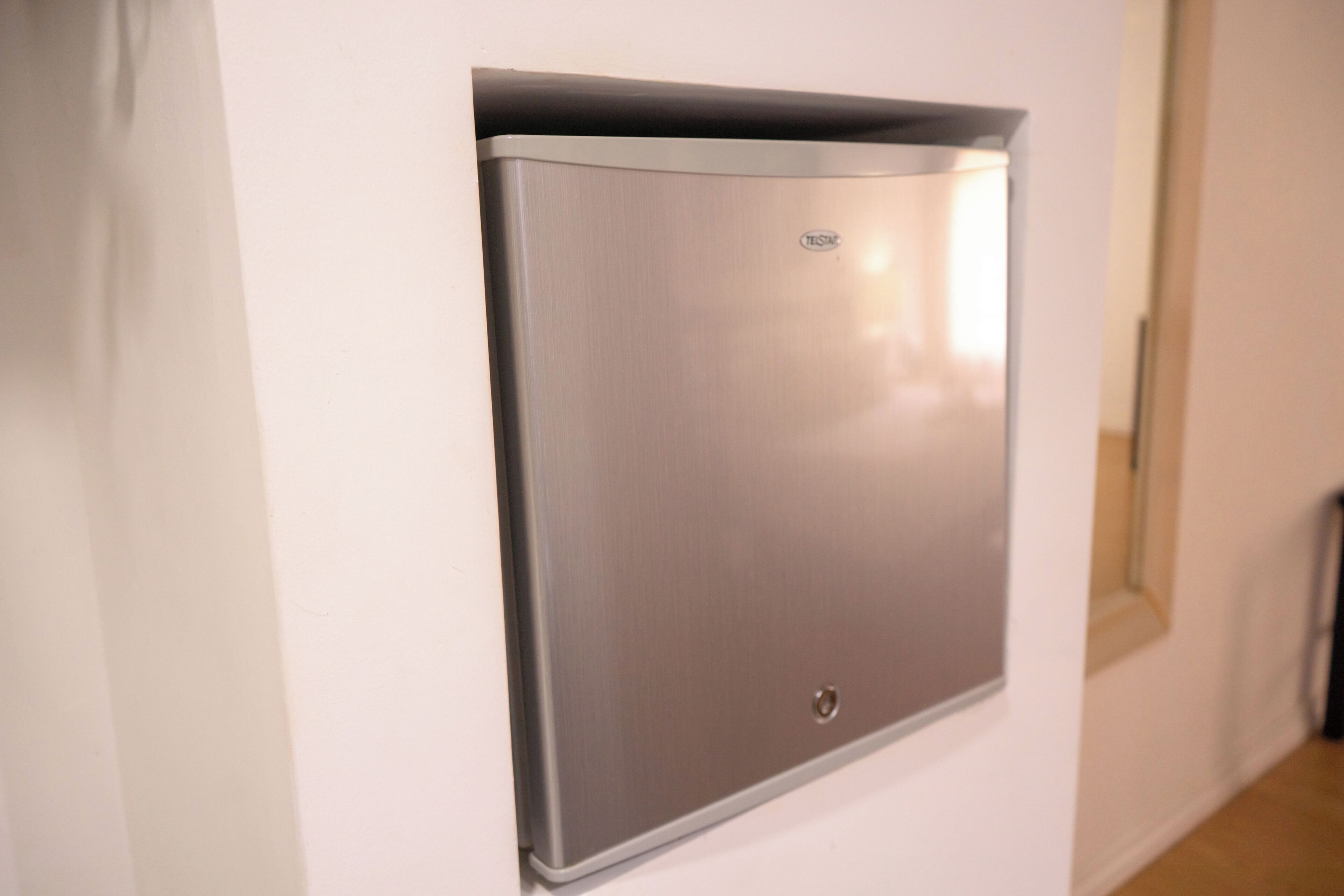Un objeto metálico fijado a la pared con un pequeño logotipo, probablemente un termostato o panel de control, en un pasillo con paredes blancas y una puerta al fondo.
