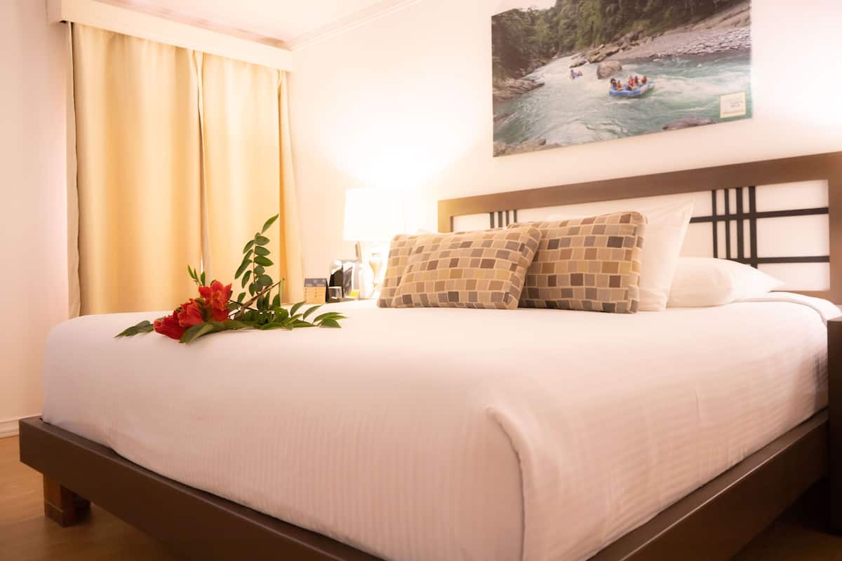 A hotel room with a bed, a painting of a river scene, a lamp, and a plant with red flowers.