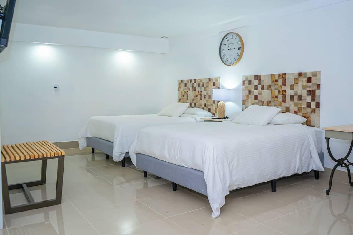 A hotel room with two beds, a wooden side table, a wall clock, and a lamp.