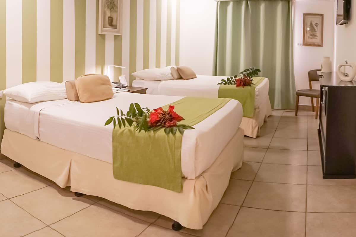 A hotel room with two beds, each with a green blanket and a red flower. There is a desk with a chair, a mirror, and a small plant. The room has a striped wall and a green curtain.