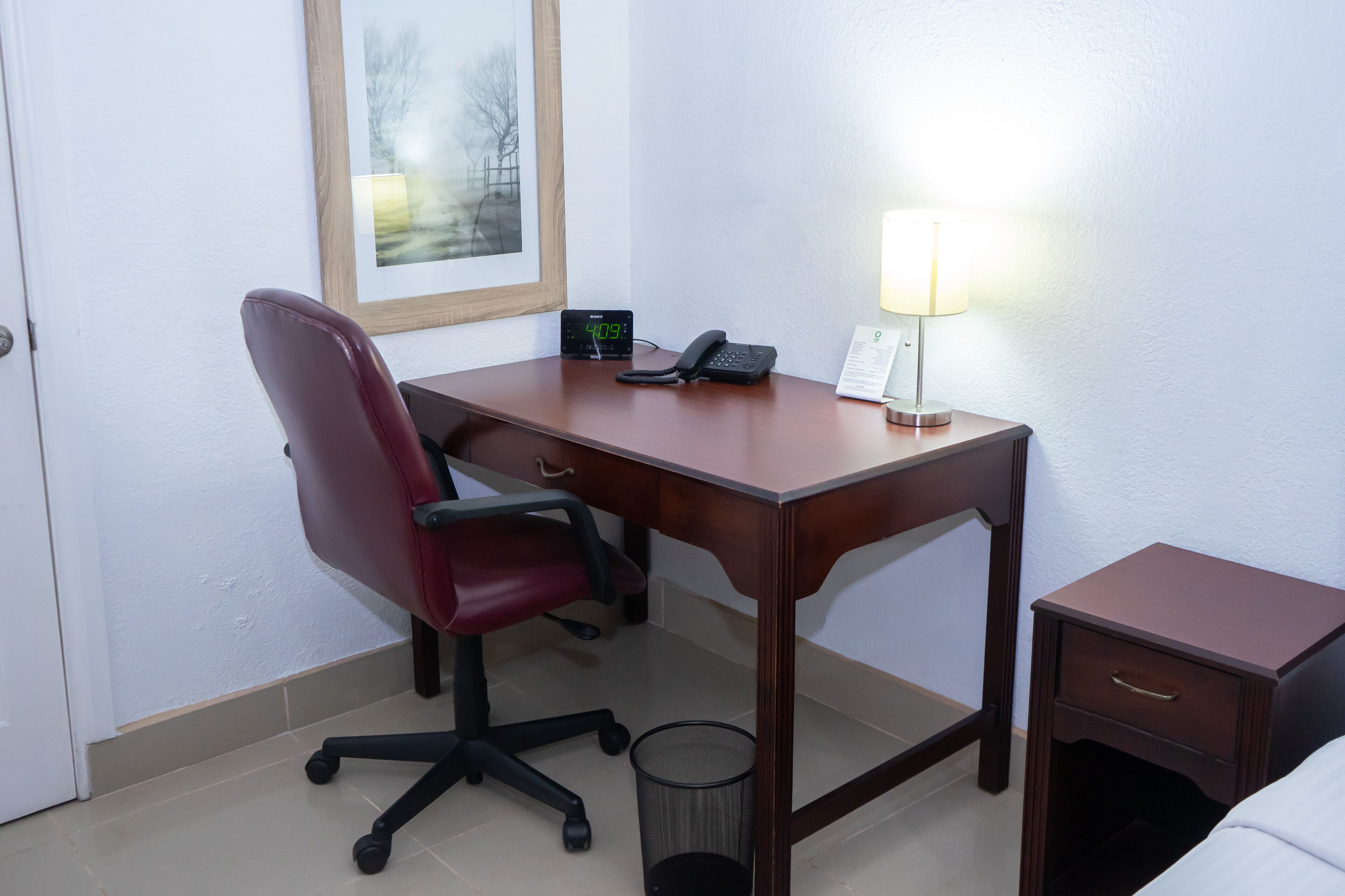 Habitación de hotel con un escritorio de madera, una silla de oficina roja, un teléfono, una lámpara y una mesita de noche pequeña.