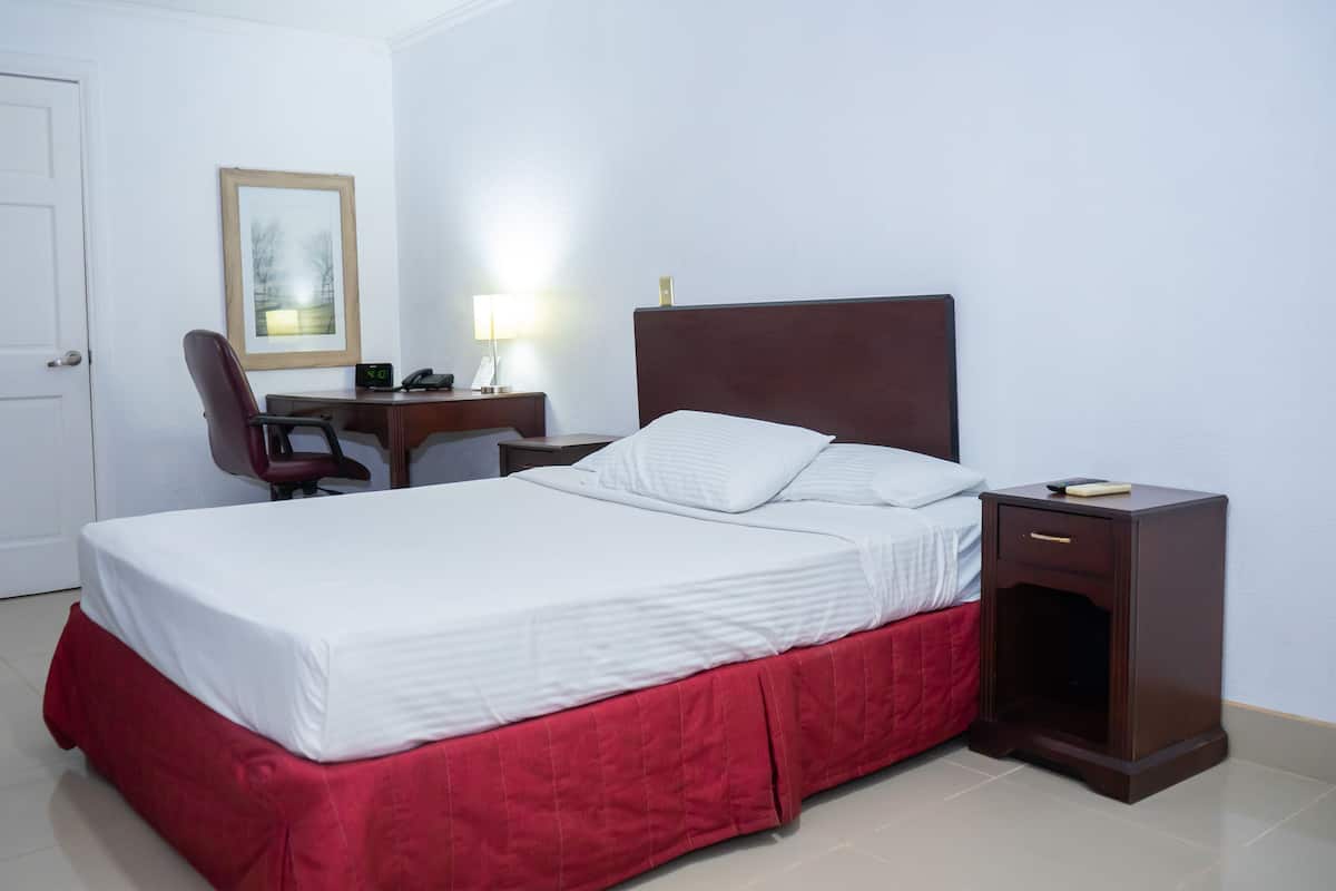 A neatly made bed with a red bed skirt, a wooden nightstand with a lamp, and a desk with a chair.