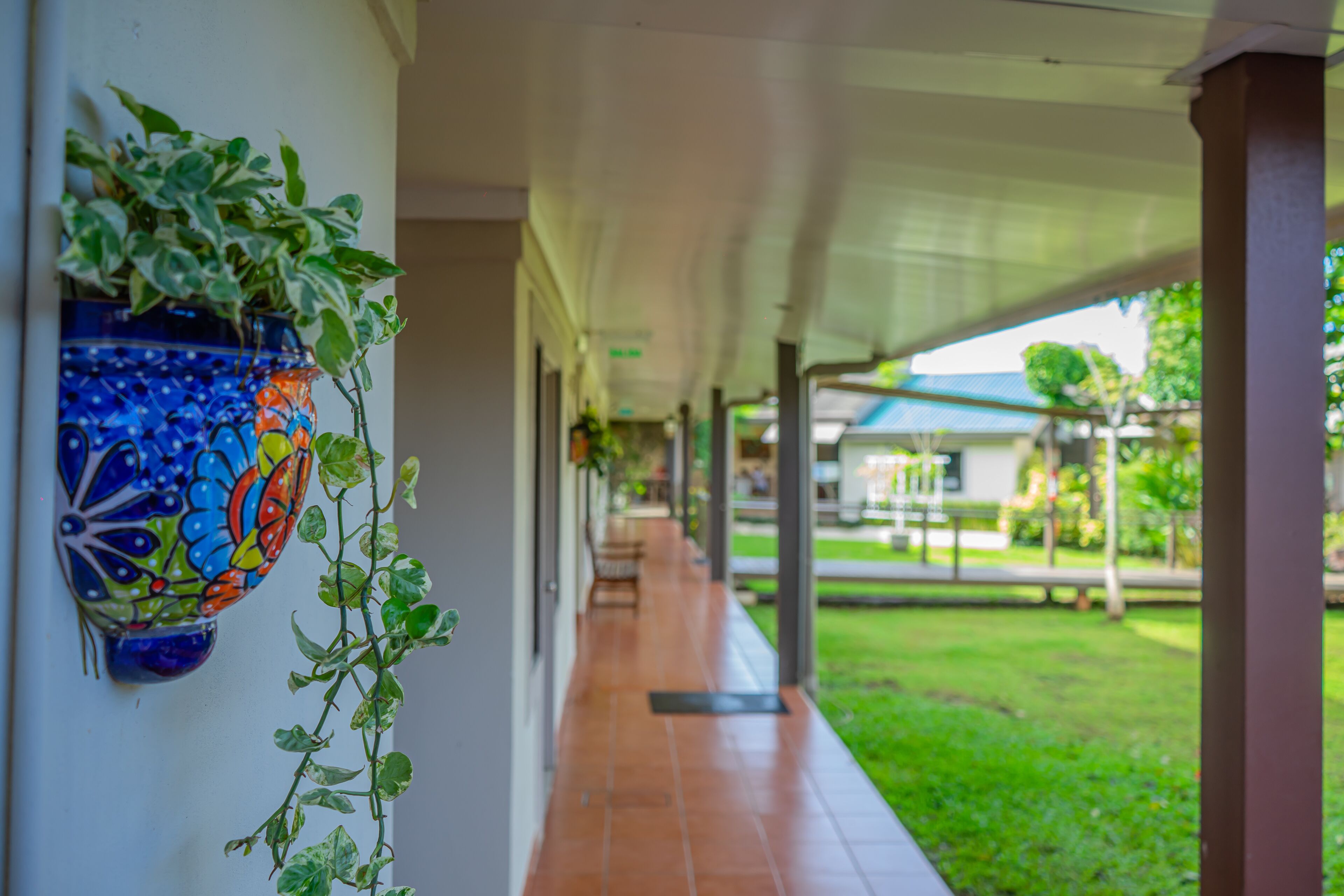 A covered corridor with a tiled floor, a decorative wall-mounted planter, and a view of a grassy area with a building in the background.