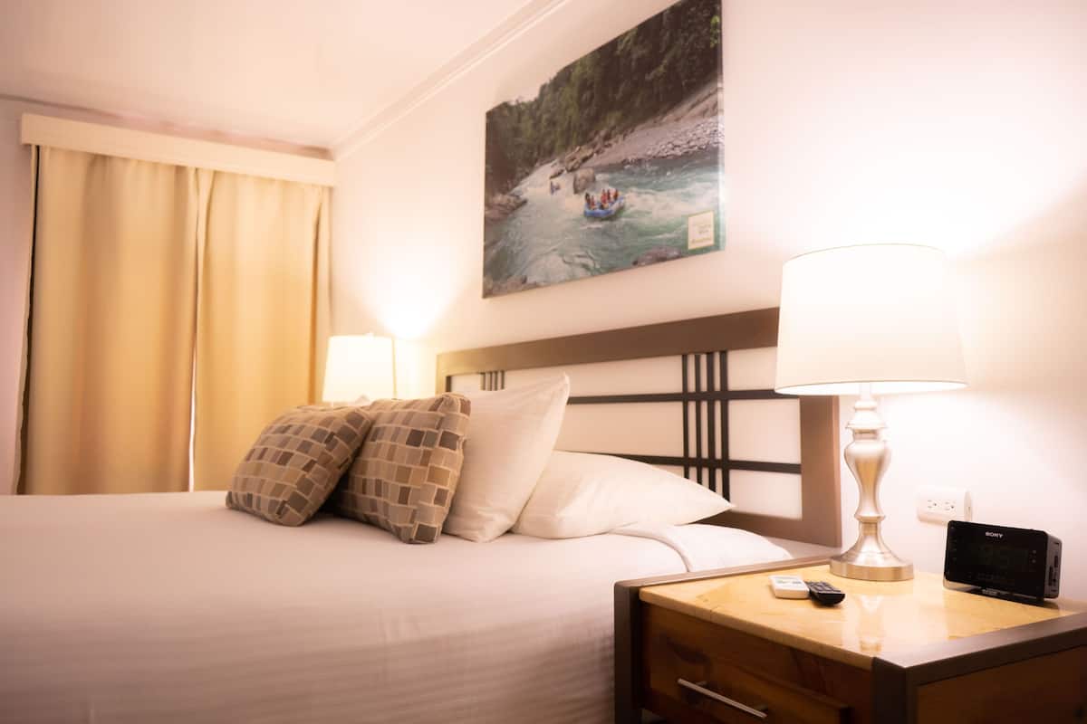 A hotel room with a bed, a nightstand with a lamp, a Sony alarm clock, and a painting of a river scene on the wall.