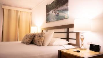 A hotel room with a bed, a nightstand with a lamp, a Sony alarm clock, and a painting of a river scene on the wall.