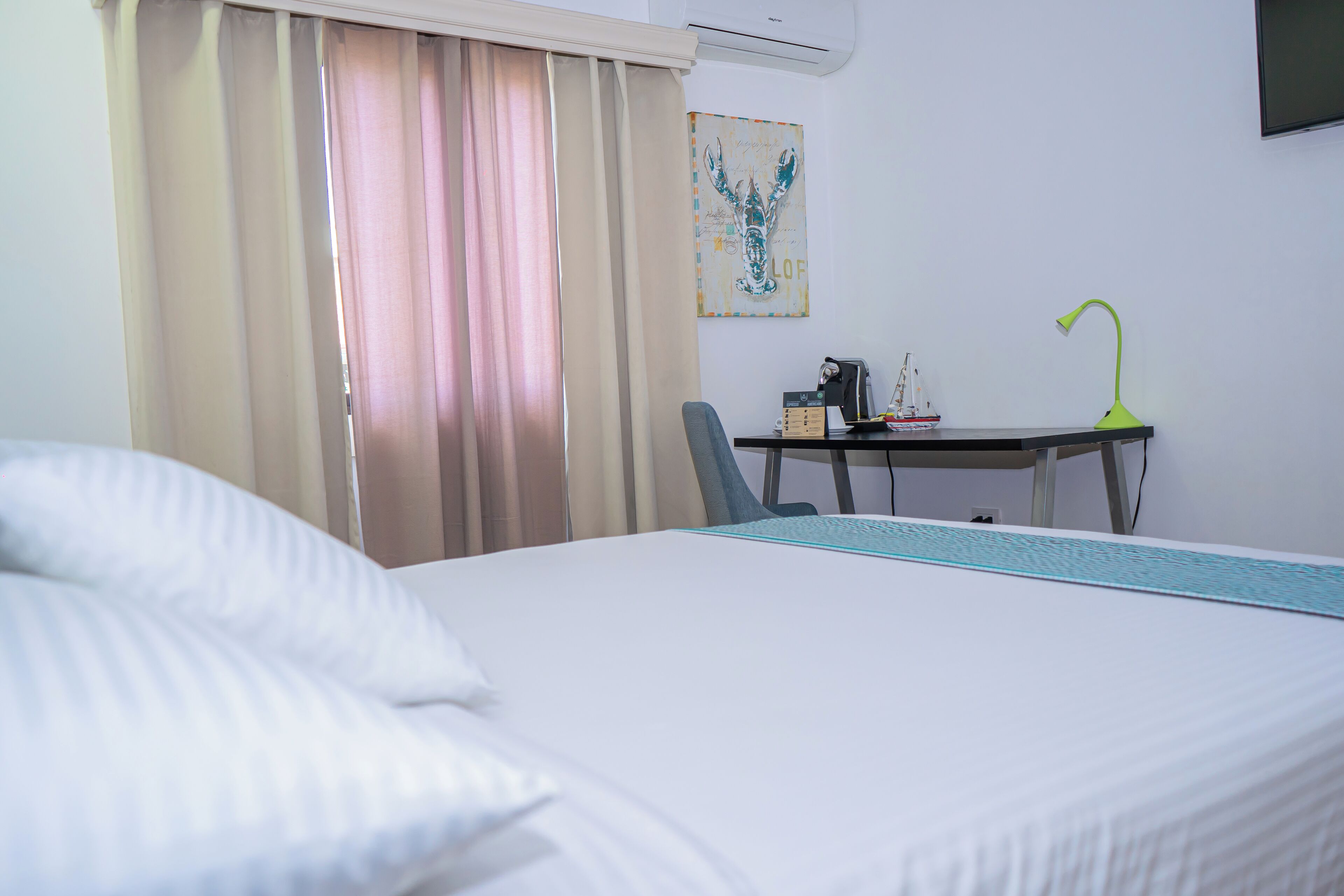 A hotel room with a bed, a desk with a lamp, a chair, and a painting on the wall.