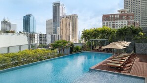 Una piscina en la azotea con sillones de descanso y sombrillas, rodeada de vegetación y con vistas a un paisaje urbano de edificios altos.