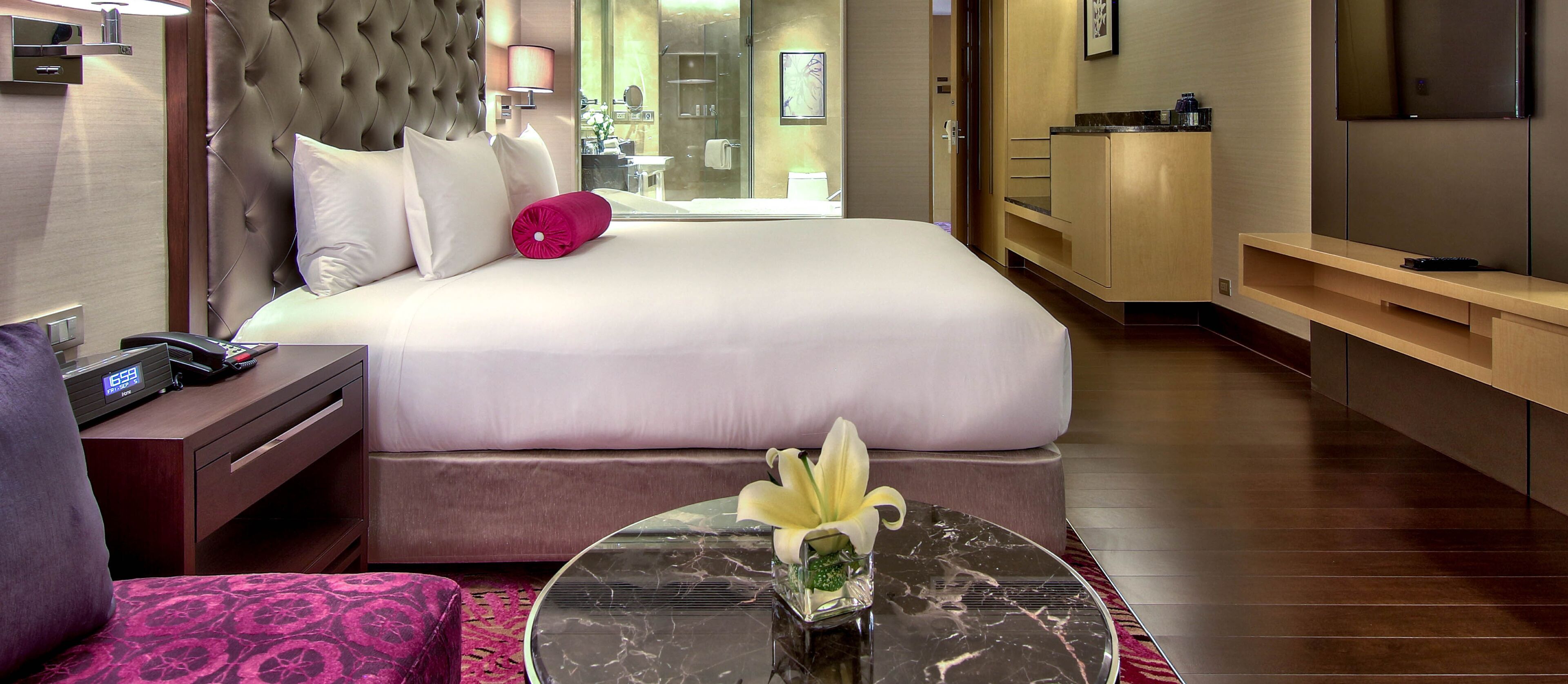A hotel room with a large bed, bedside table, lamp, and a small table with a flower.