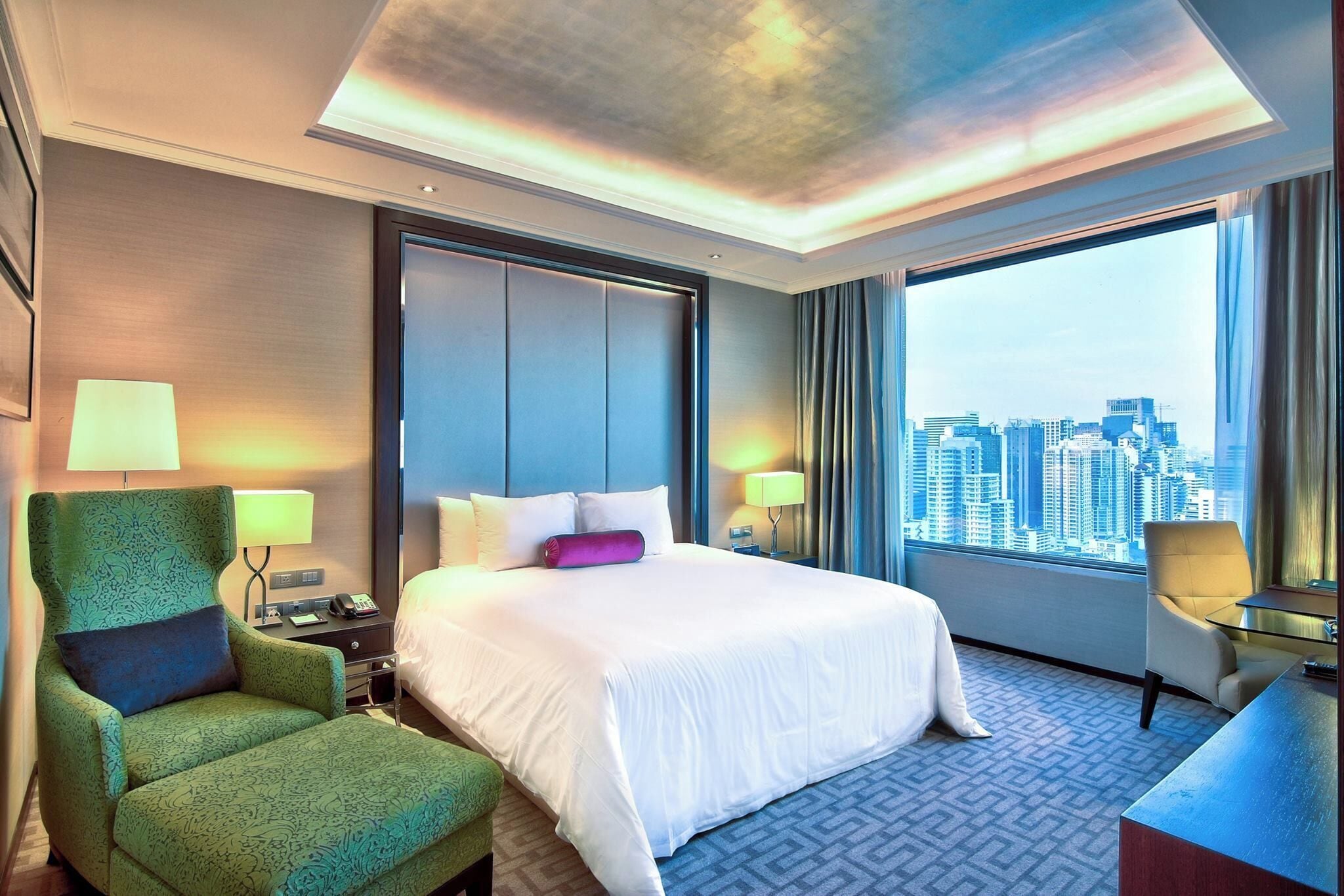 A hotel room with a large bed, a green armchair, a desk, and a city view through the window.