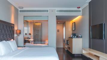 Habitación de hotel moderna con una cama grande, televisión, baño con puerta de vidrio y kitchenette.