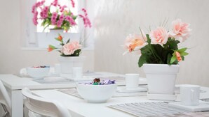 Una mesa de comedor con sillas blancas, un cuenco blanco con aperitivos y dos plantas en macetas blancas con flores.