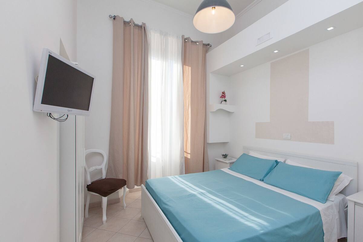 A bedroom with a bed, a TV mounted on the wall, a chair, and a bedside table.