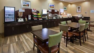 A breakfast area with a refrigerator, coffee machine, and various food items. There are tables and chairs for dining.