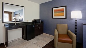 A hotel room with a kitchen area, a chair, a lamp, and a painting on the wall.