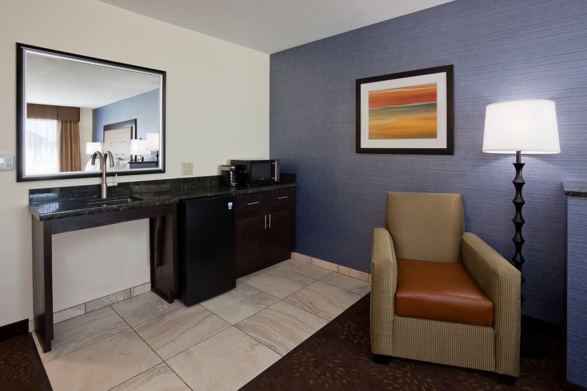 A hotel room with a kitchen area, a chair, a lamp, and a painting on the wall.