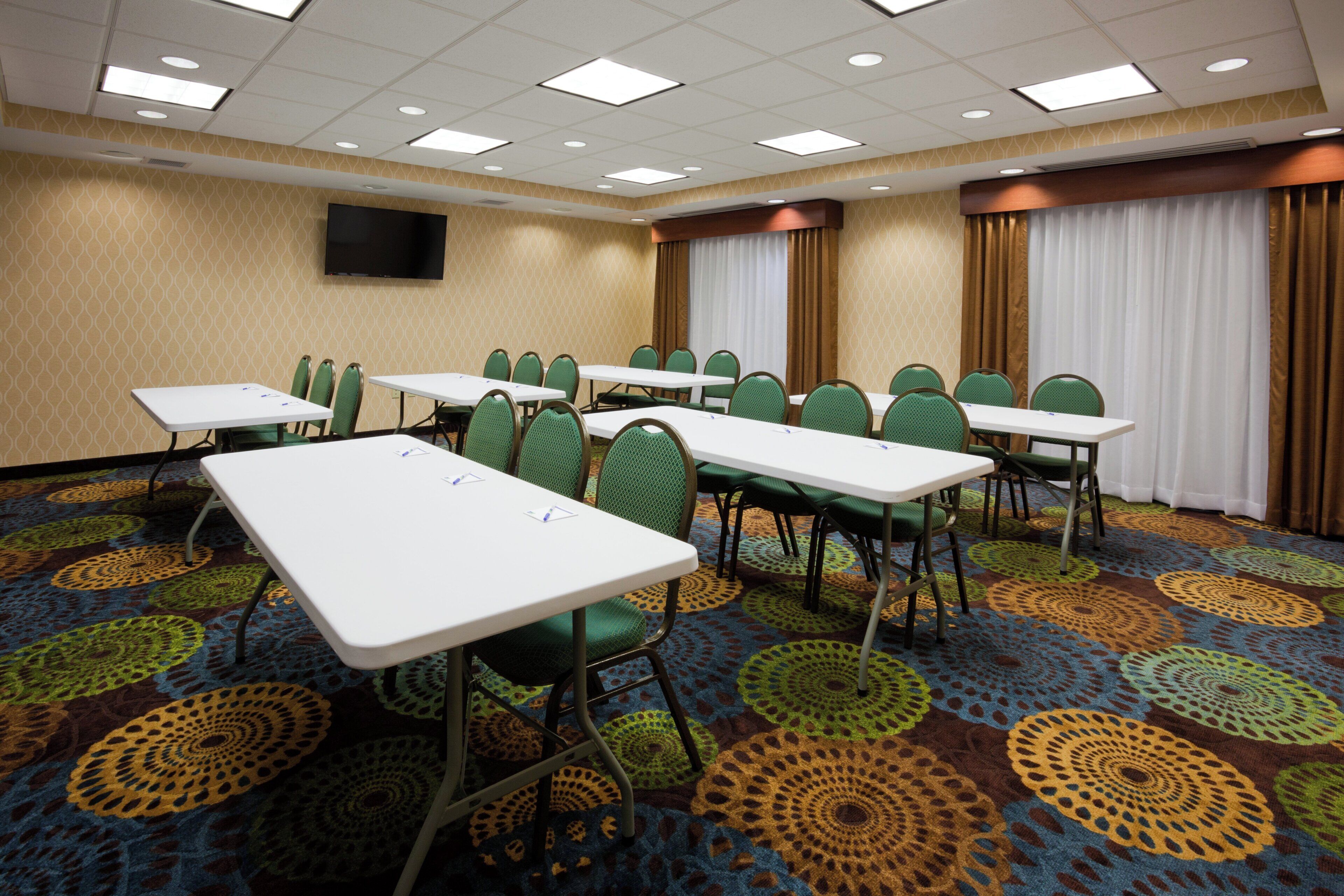 A conference room with multiple tables and chairs, a flat-screen TV mounted on the wall, and patterned carpet.