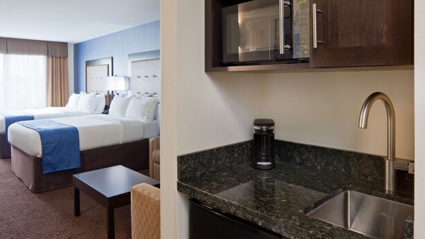 A hotel room with two beds, a small table, a coffee maker, a microwave, and a sink.