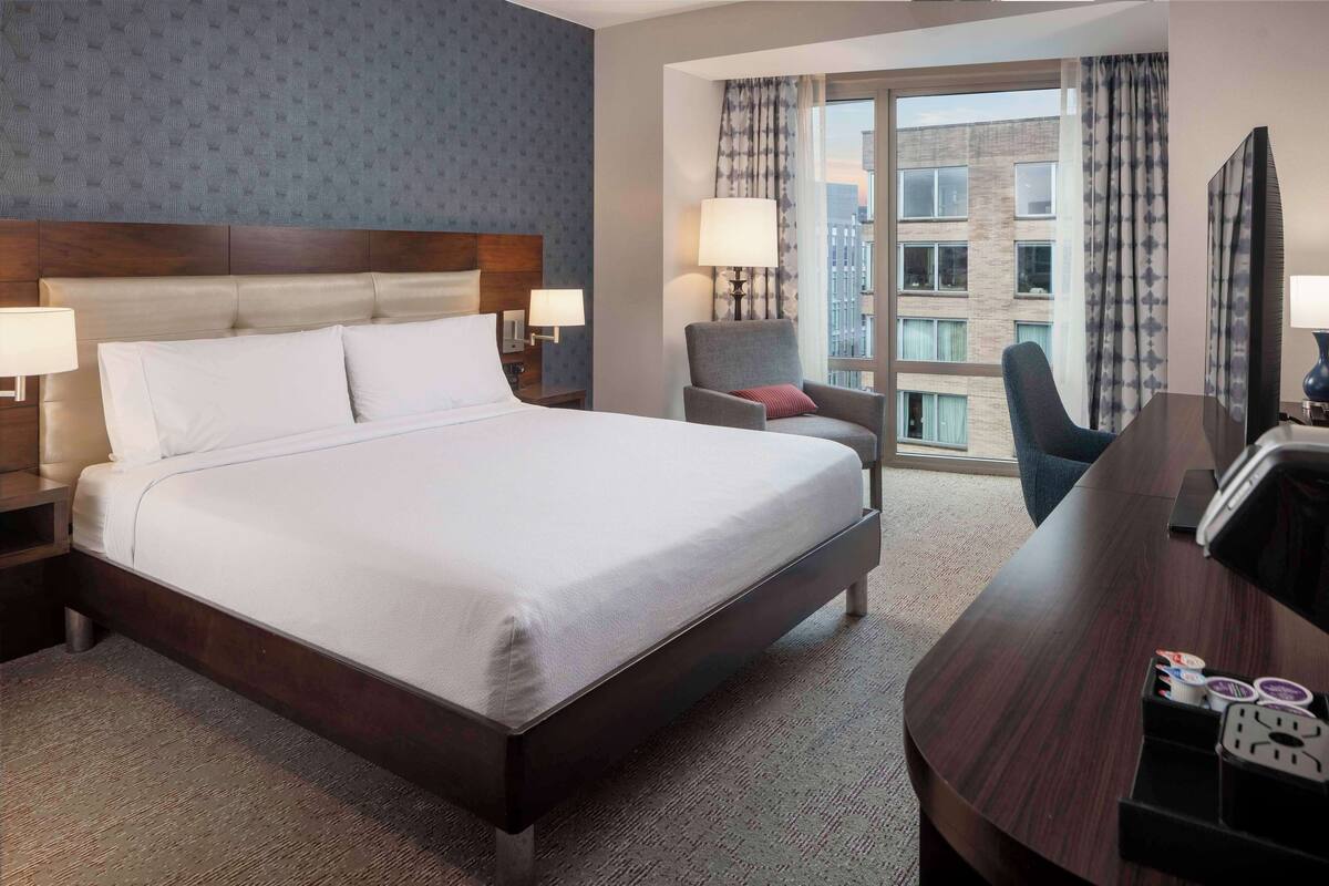 A hotel room with a large bed, a desk, a chair, and a window with a view of buildings.