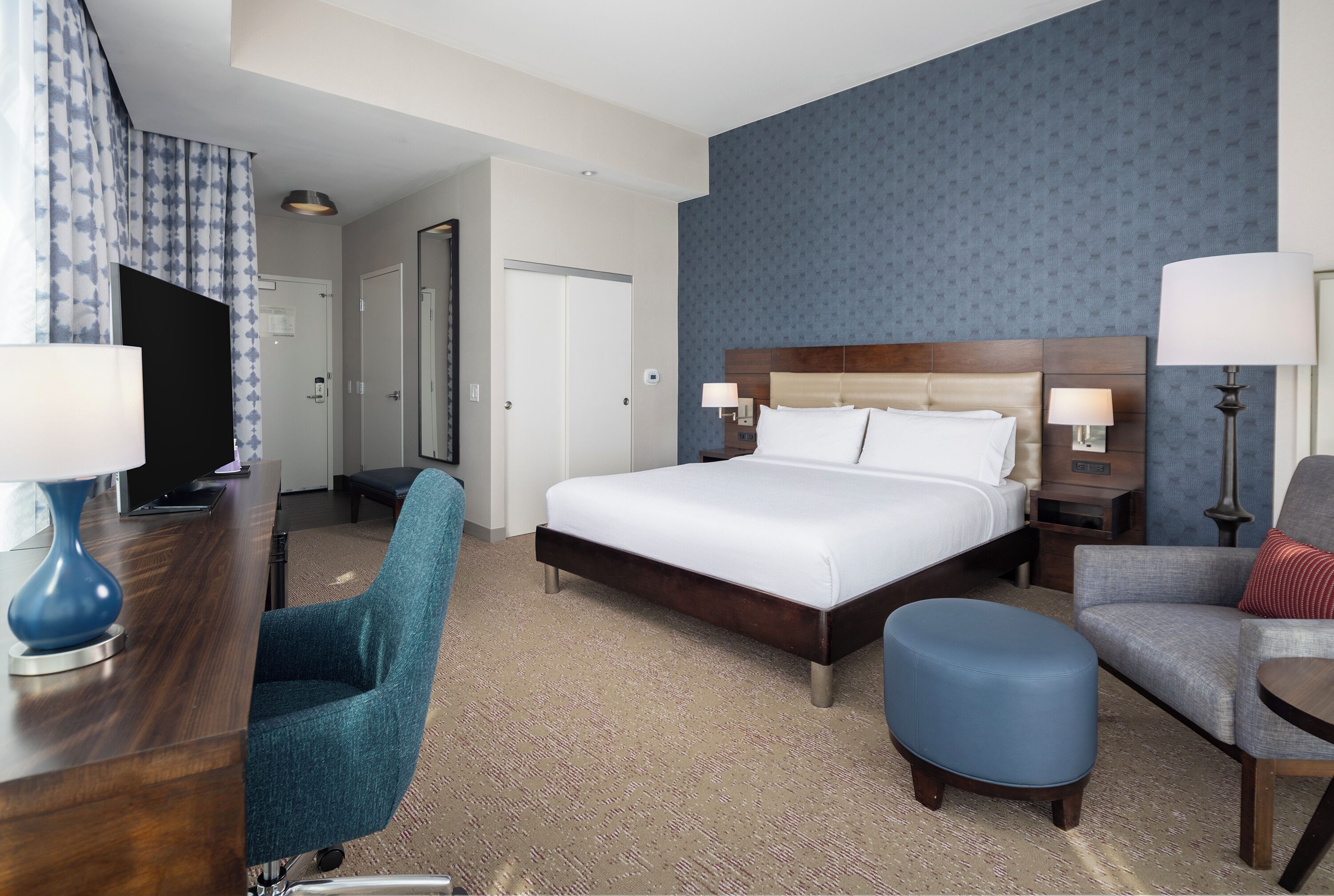 A hotel room with a large bed, a desk with a computer, a chair, a blue ottoman, and a lamp.