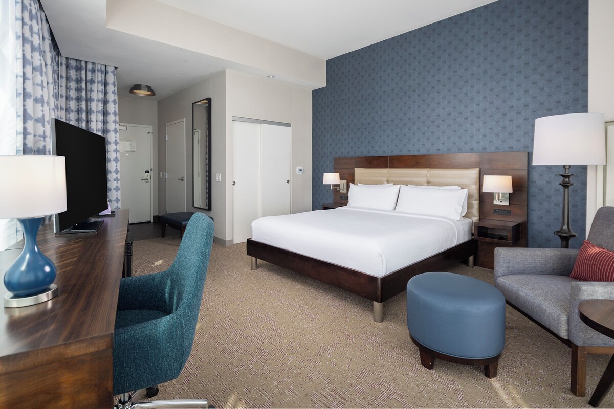 A hotel room with a large bed, a desk with a computer, a chair, a blue ottoman, and a lamp.
