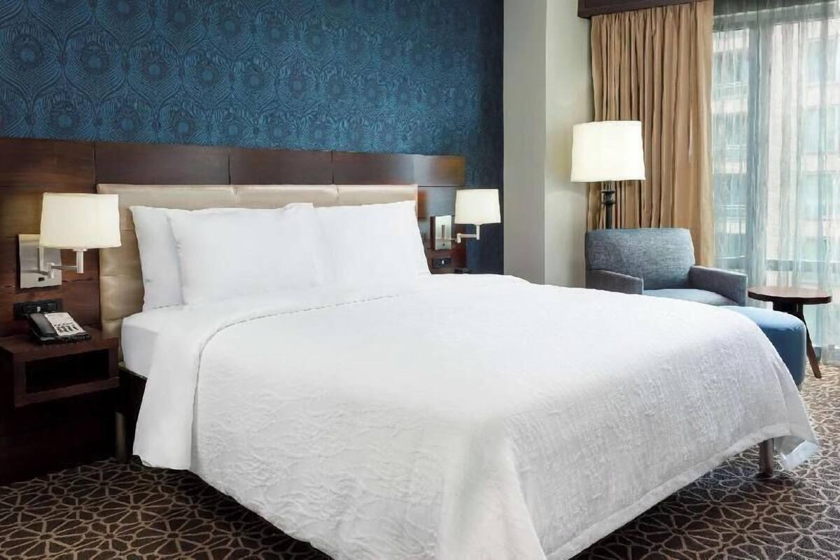 A hotel room with a large bed, two bedside tables with lamps, a chair, and a window with curtains.