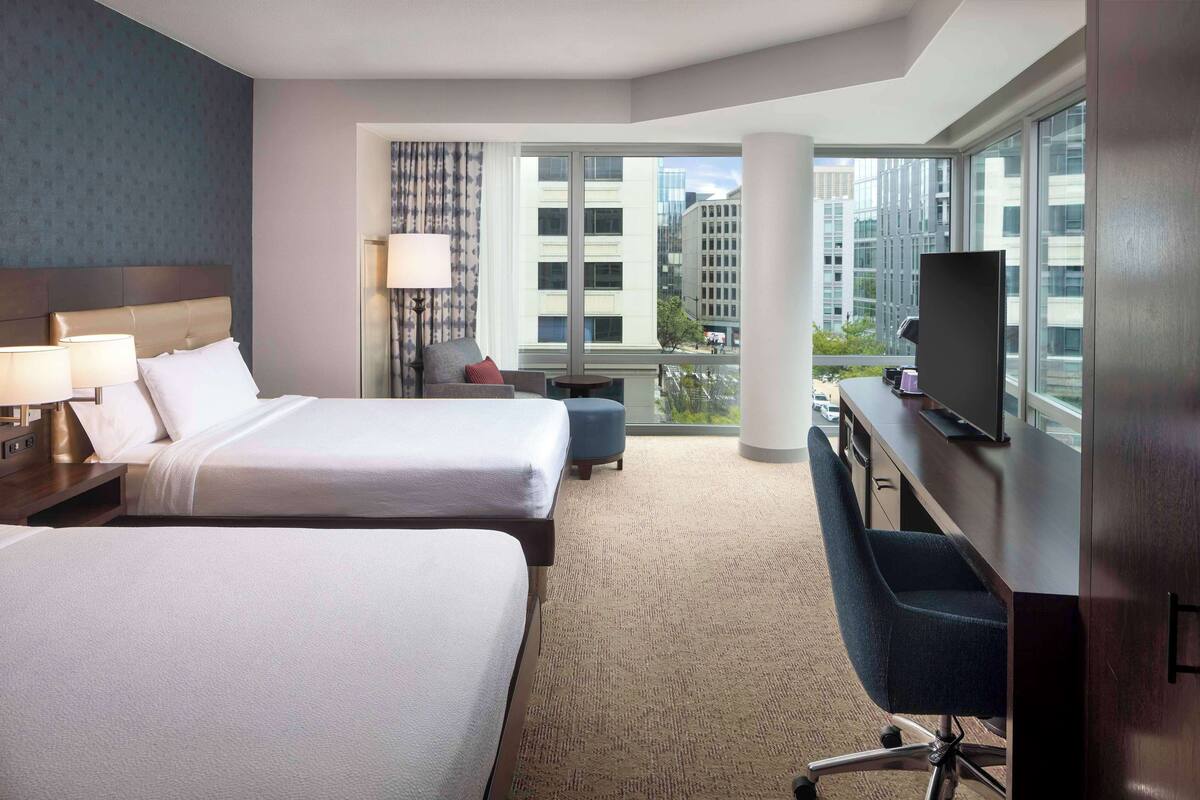 A hotel room with two beds, a desk with a TV, a chair, and a large window offering a city view.