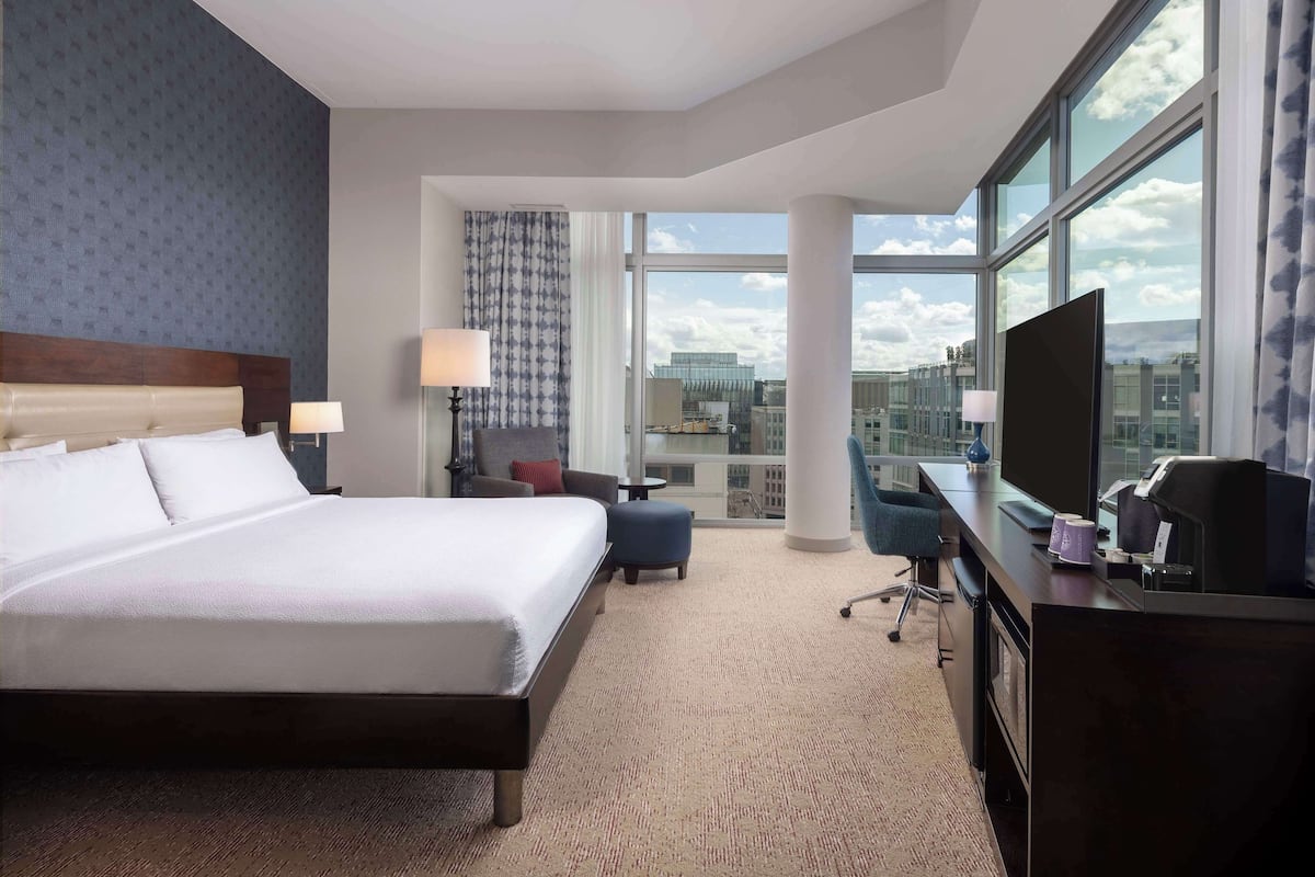 A hotel room with a large bed, a desk with a computer, a TV, and a view of the city through large windows.
