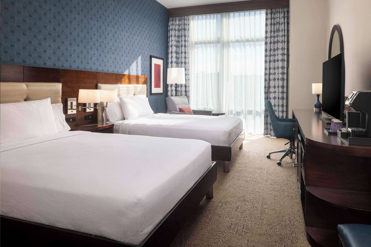 A hotel room with two beds, a desk, a chair, a TV, and a large window with curtains.
